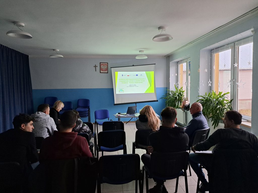 Zdjęcie przedstawia uczestników spotkania siedzących na krzesłach. Na przeciwko jest widoczna wyświetlona prezentacja