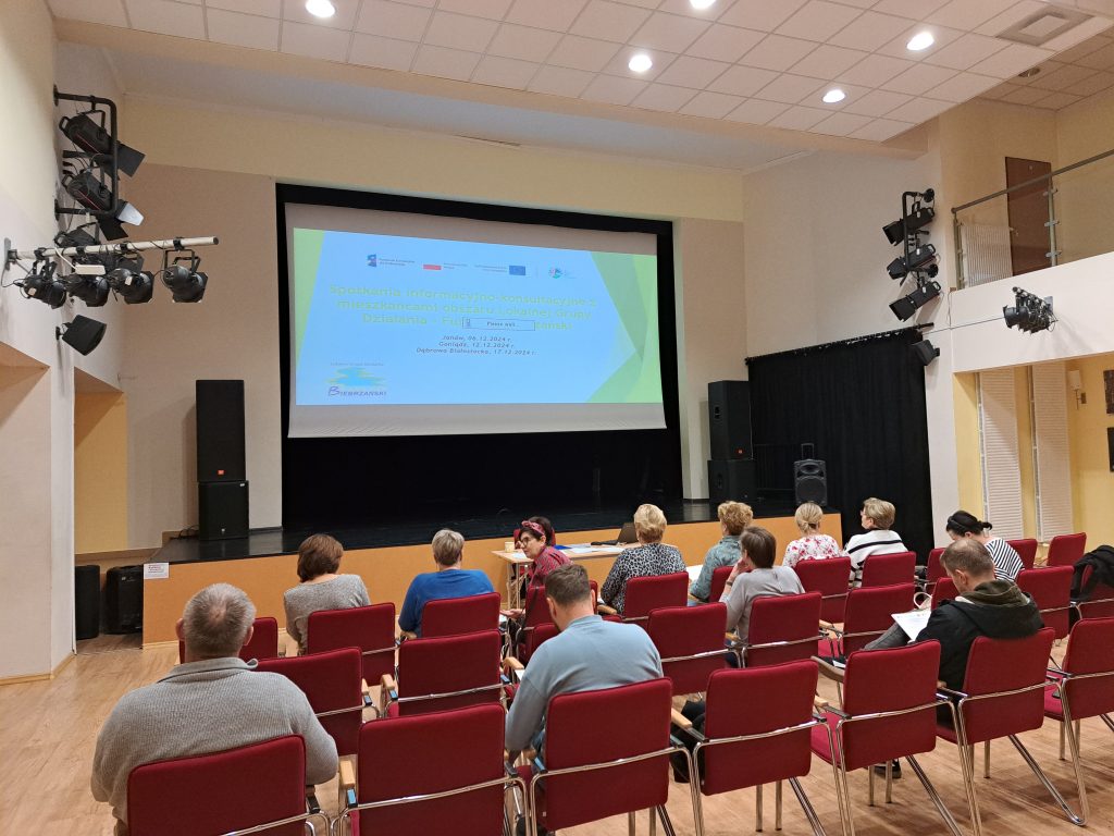 Zdjęcie przedstawia uczestników spotkania siedzących na krzesłach. Na przeciwko jest widoczna wyświetlona prezentacja