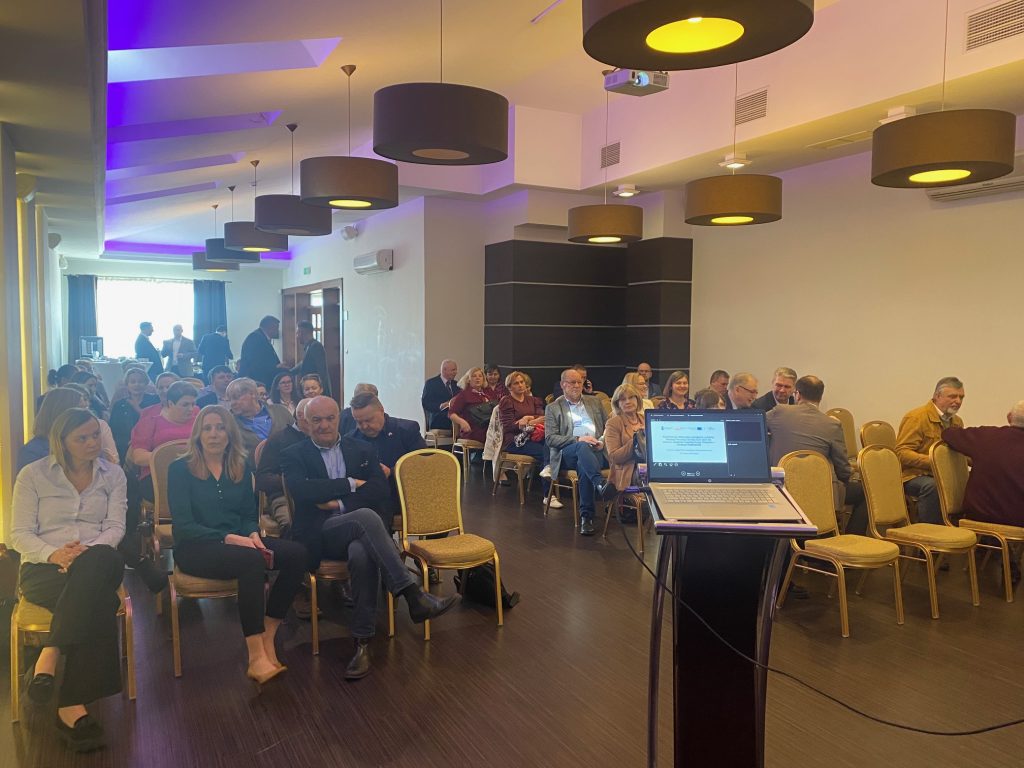 Zdjęcie przedstawia uczestników konferencji siedzących w sali w rozmawiających podczas przerwy. Część uczestników stoi w głębi sali.