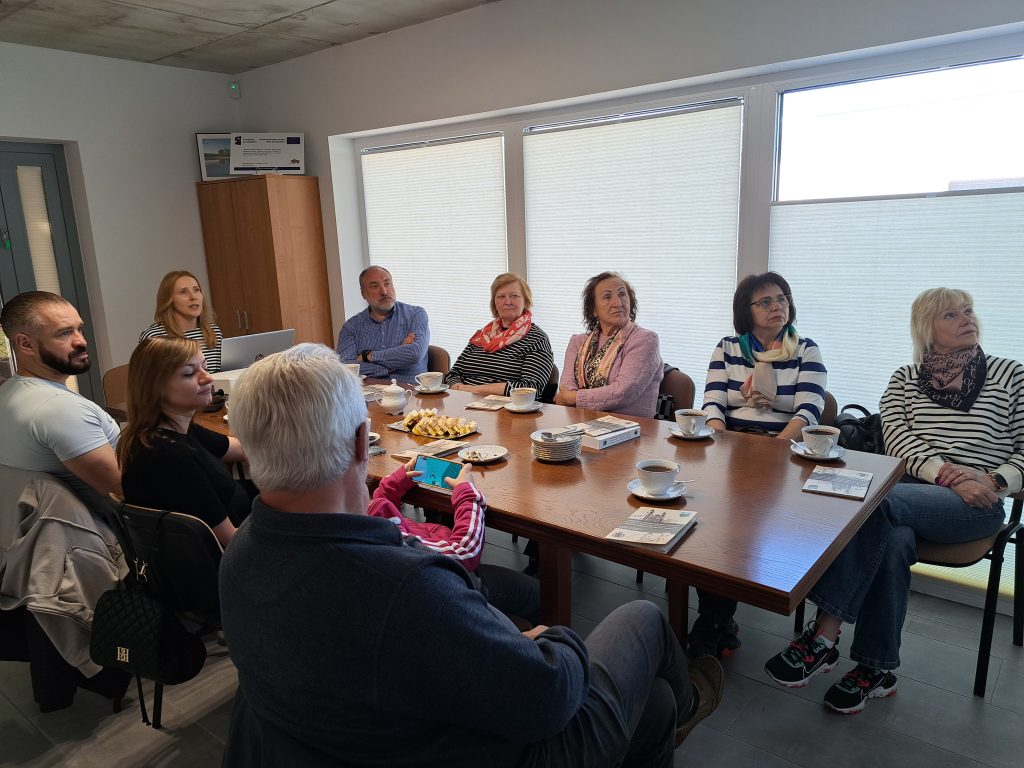 Zdjęcie przedstawia uczestników spotkania, którzy siedzą przy stole i oglądają wyświetlaną prezentację. Prezentację przedstawia pracownica biura Lokalnej Grupy Działania.