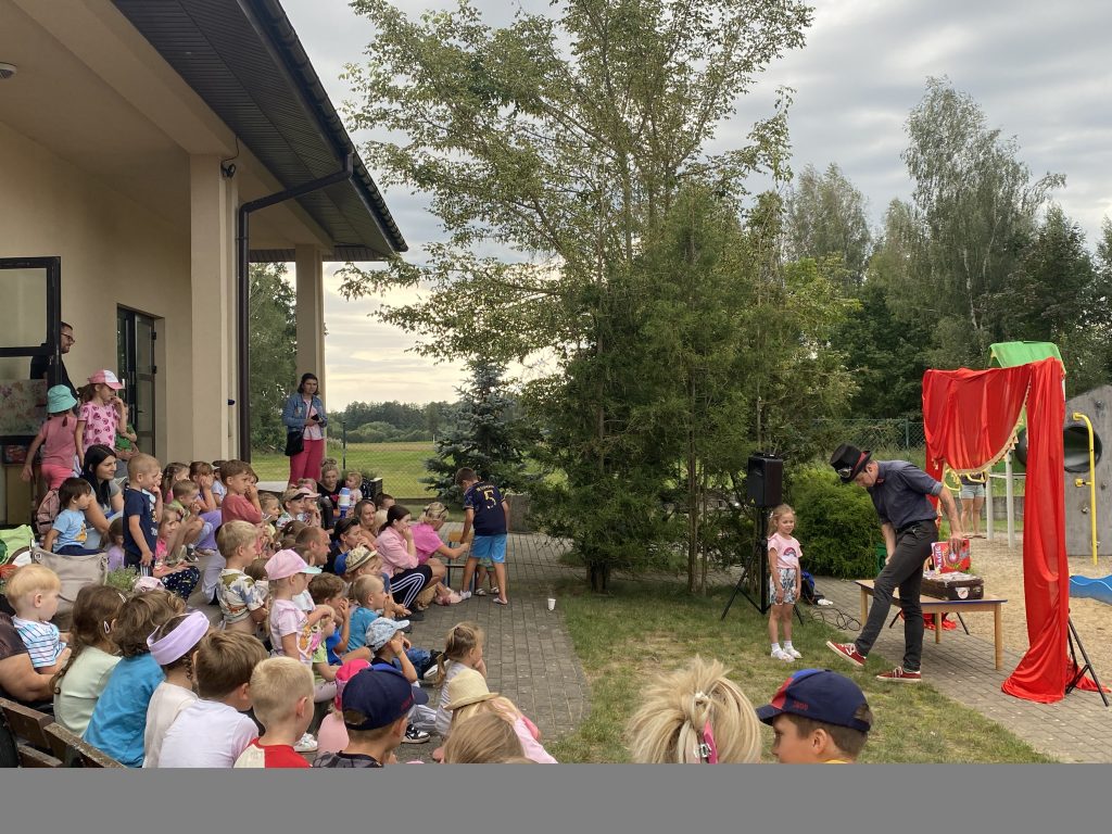 Zdjęcie przedstawia dzieci w wieku przedszkolnym, które siedzą na schodach przed budyniem świetlicy wiejskiej. Naprzeciw dzieci swój pokaz przedstawia komik, który ubrany jest w czarny strój i ma na głowie cylinder.